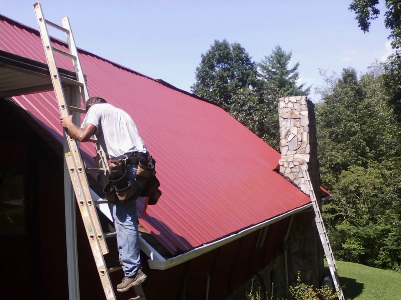 Roofer on a ladder beside a red metal roof and stone chimney — Soffit & Fascia Repair by Roofing Brothers in Summit Lake
